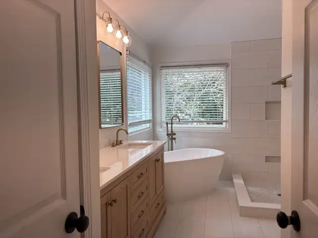 Light tile bathroom floor running from a wood vanity past a freestanding tub to the shower curb