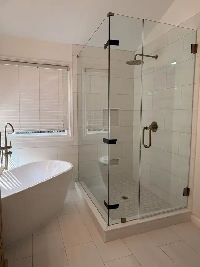 Frameless glass shower enclosure with brass handle next to a freestanding tub